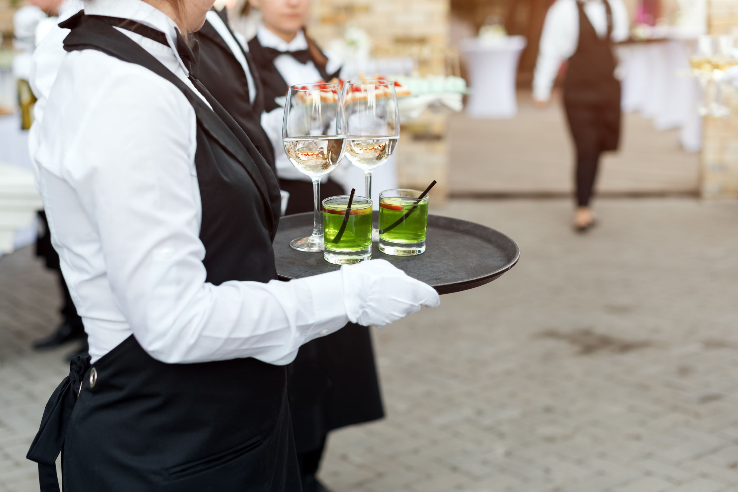 Midsection of professional waiters in uniform serving wine, cocktails and snacks during buffet catering party, festive event or wedding. Full glasses of wine on tray. Outdoor party catering service.
