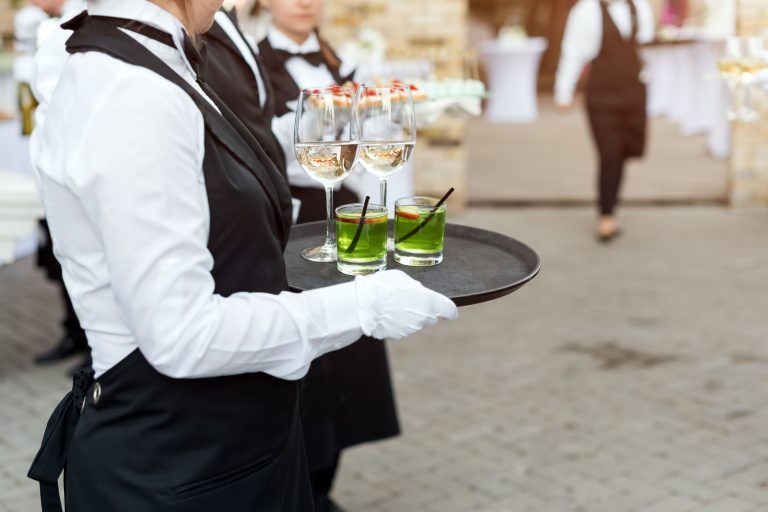 Midsection of professional waiters in uniform serving wine, cocktails and snacks during buffet catering party, festive event or wedding. Full glasses of wine on tray. Outdoor party catering service.